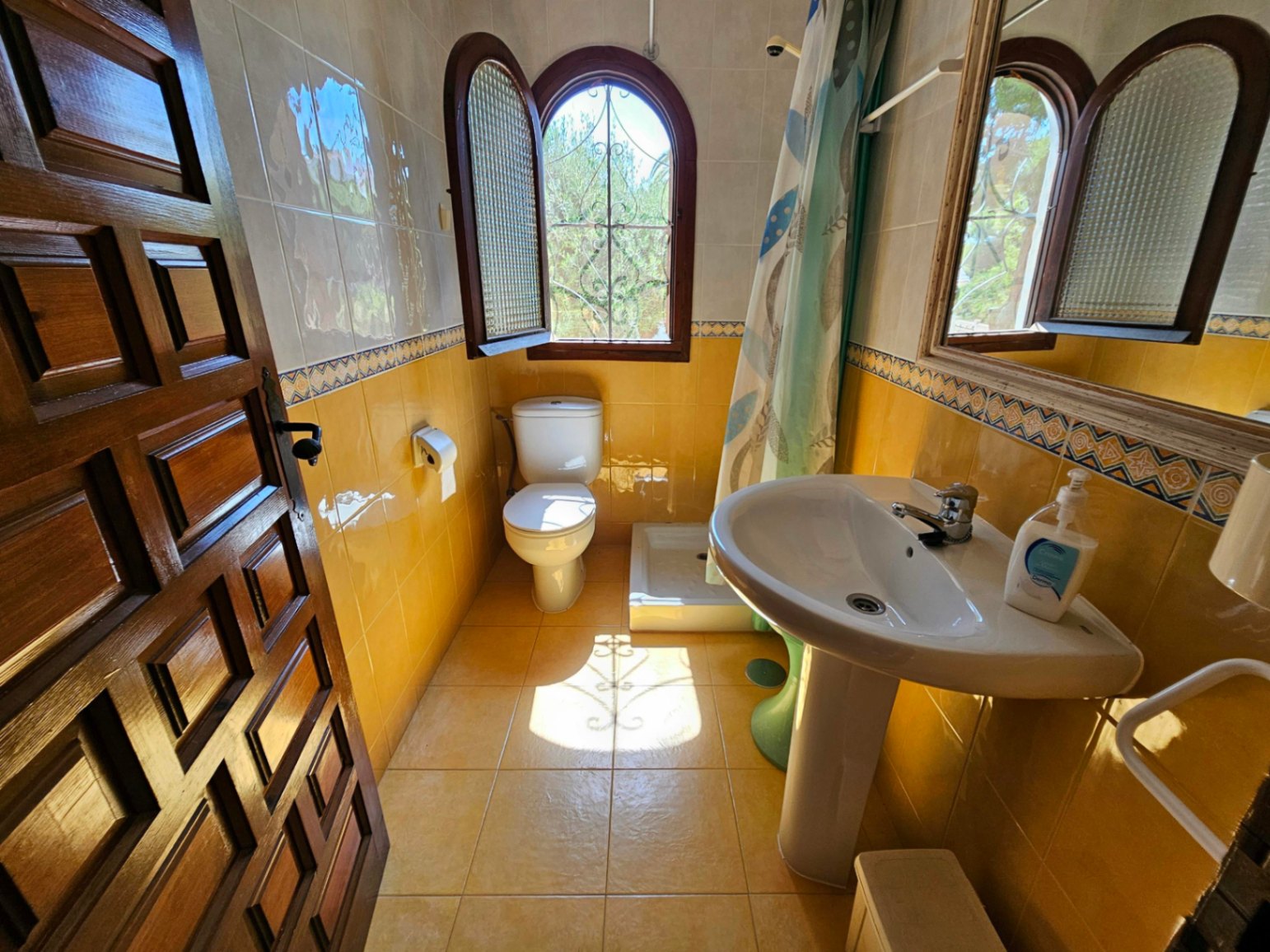 Traditionelles Badezimmer mit gelben Wandfliesen, weißem Säulenwaschbecken und einem gewölbten Holzfenster mit Ziergitter.