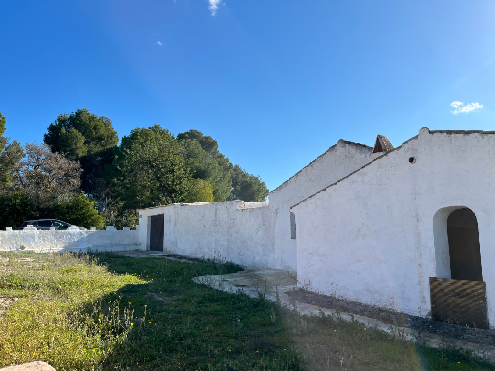 Außenansicht einer traditionellen, weiß getünchten Finca in Jávea mit Rundbogeneingang, Steinmauern und mediterraner Vegetation unter blauem Himmel.