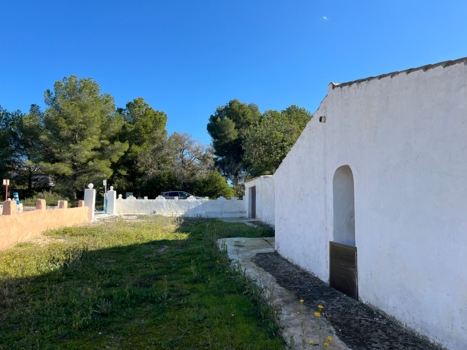 Außenansicht einer traditionellen weißen Finca in Jávea mit Rundbogennische und Naturgarten, umgeben von Pinien und einer weißen Grundstücksmauer.