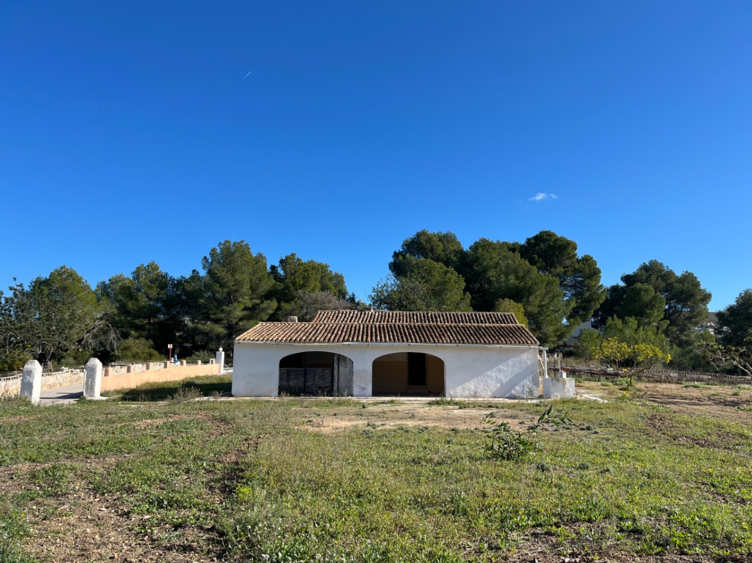 Außenansicht einer traditionellen weißen Finca mit charakteristischer Rundbogen-Terrasse und Ziegeldach auf einem großen Naturgrundstück mit Pinien.
