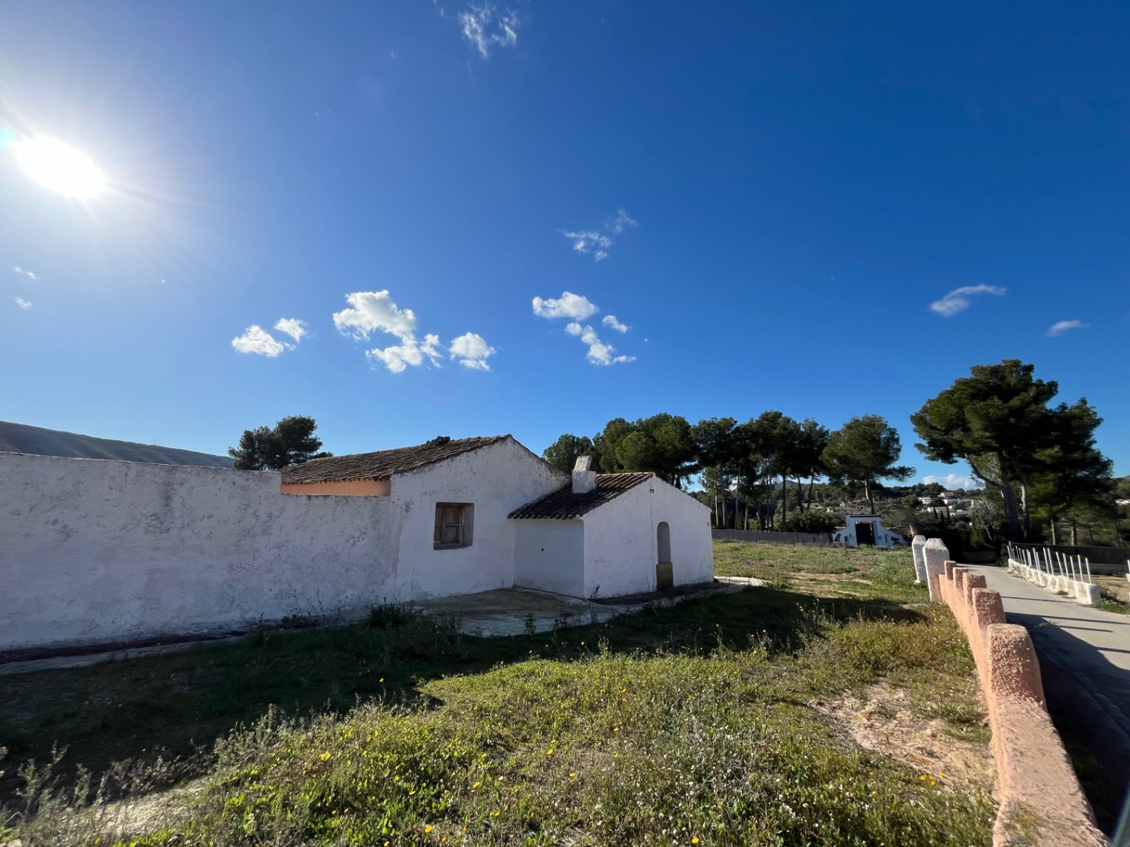 Außenansicht einer traditionellen weißen Finca mit Ziegeldach auf einem Naturgrundstück mit Pinien und klarem blauem Himmel.