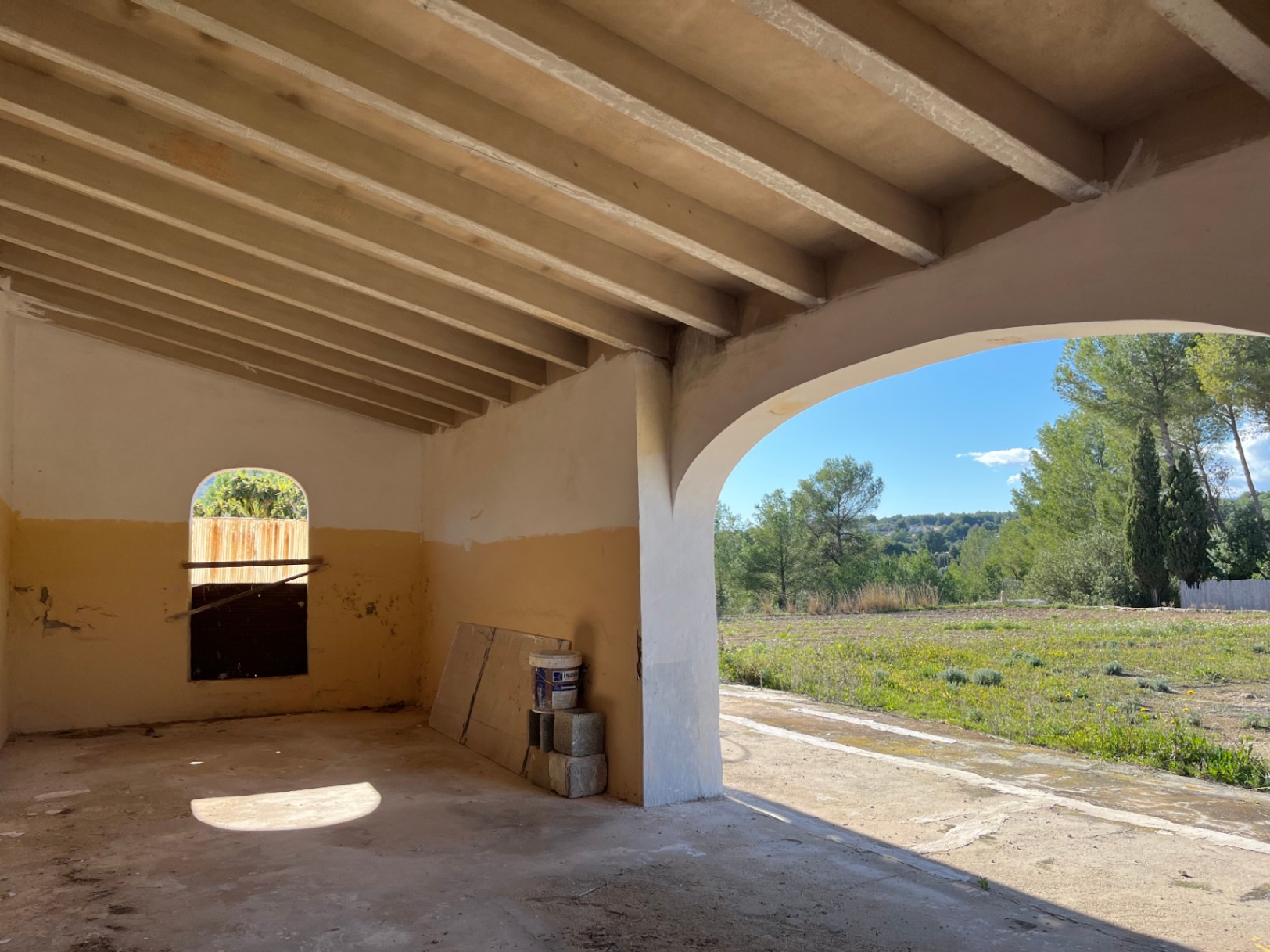 Renovierungsbedürftige Veranda mit Sichtbalkendecke und großem Rundbogen, der den Blick auf die umliegenden Felder und Pinien freigibt.