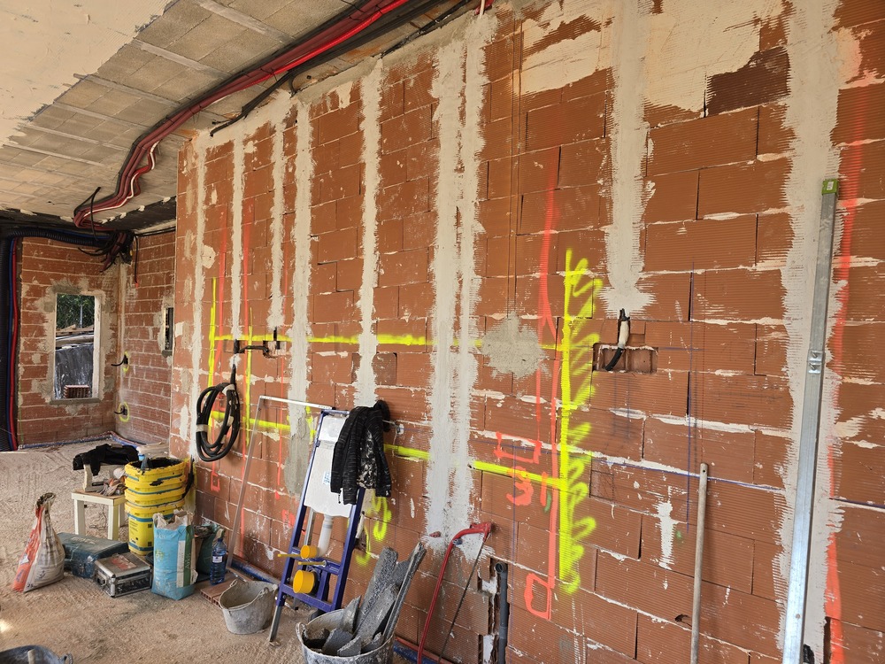 Bauphase mit einer roten Ziegelwand, fluoreszierenden Markierungen für die Elektroplanung und offen verlegten Leitungen an der Decke.