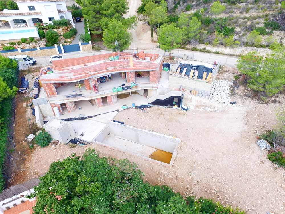 Luftaufnahme einer im Bau befindlichen Villa mit rotem Mauerwerk, Betonstrukturen und einem ausgehobenen Poolbereich in Hanglage.