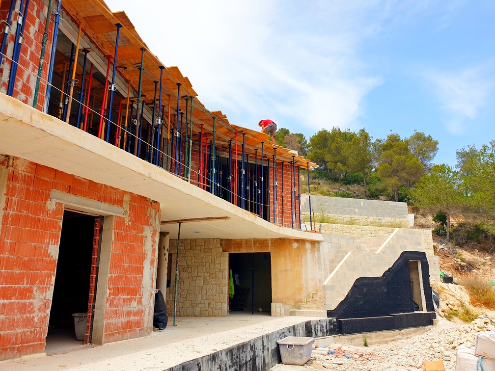 Moderne Villa im Bau mit roten Ziegelwänden, Betonplatten und Metallstützen. Die Baustelle zeigt eine Natursteinwand und eine Außentreppe aus Betonblöcken.