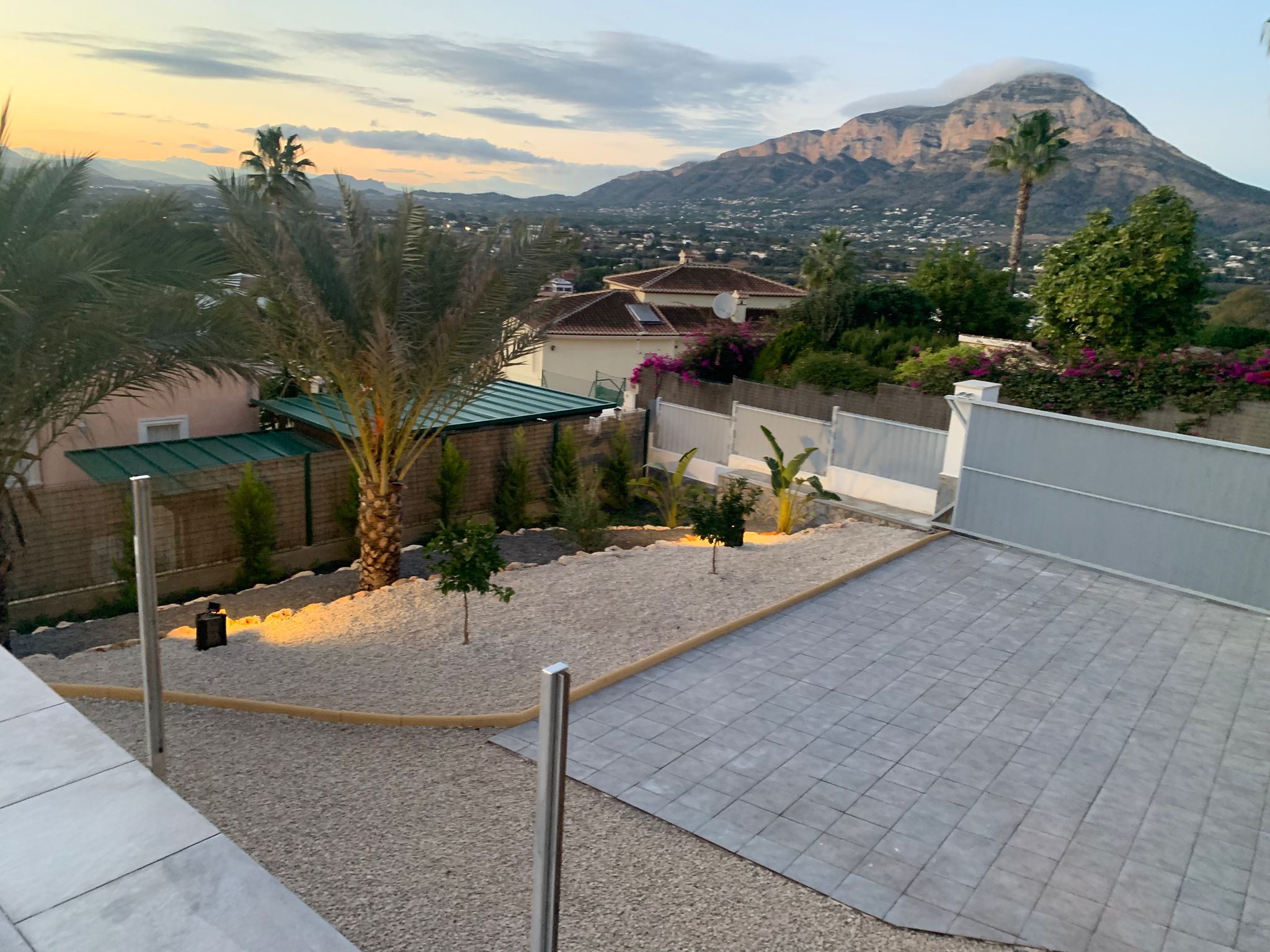 Erhöhter Blick auf einen angelegten Garten mit Kies, Palmen und gepflasterter Einfahrt, inklusive Panoramablick auf die Berge bei Sonnenuntergang.