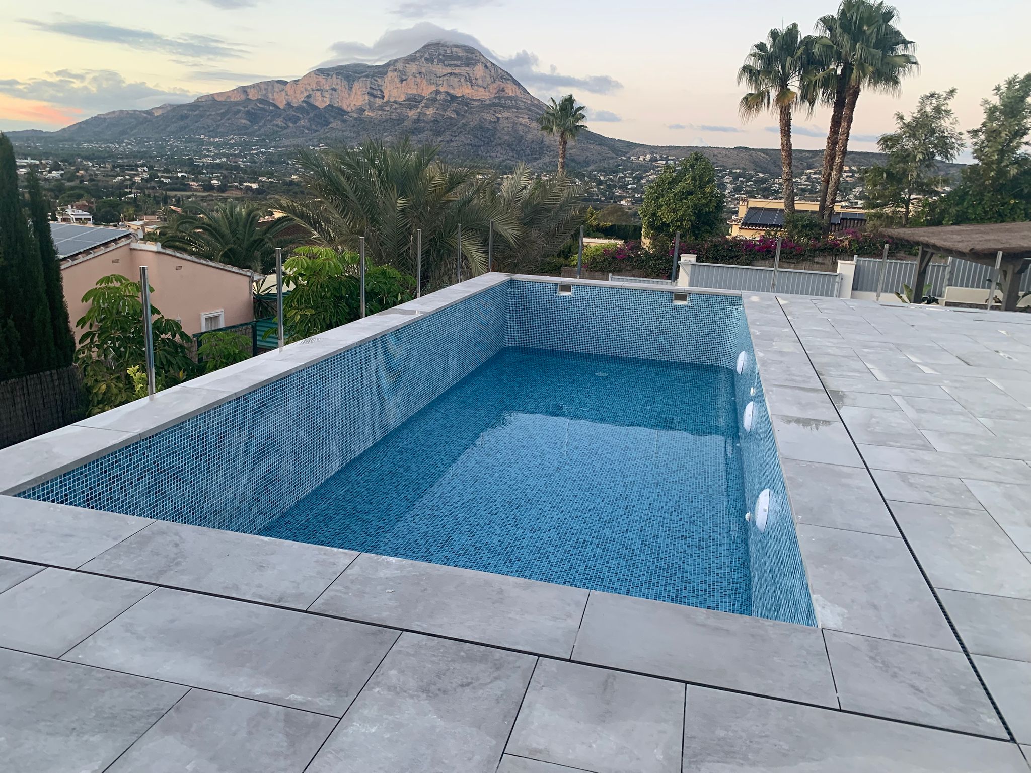 Erhöhter rechteckiger Pool mit blauen Mosaikfliesen und grauer Steinterrasse, Glasgeländer und Panoramablick auf den Berg Montgó.