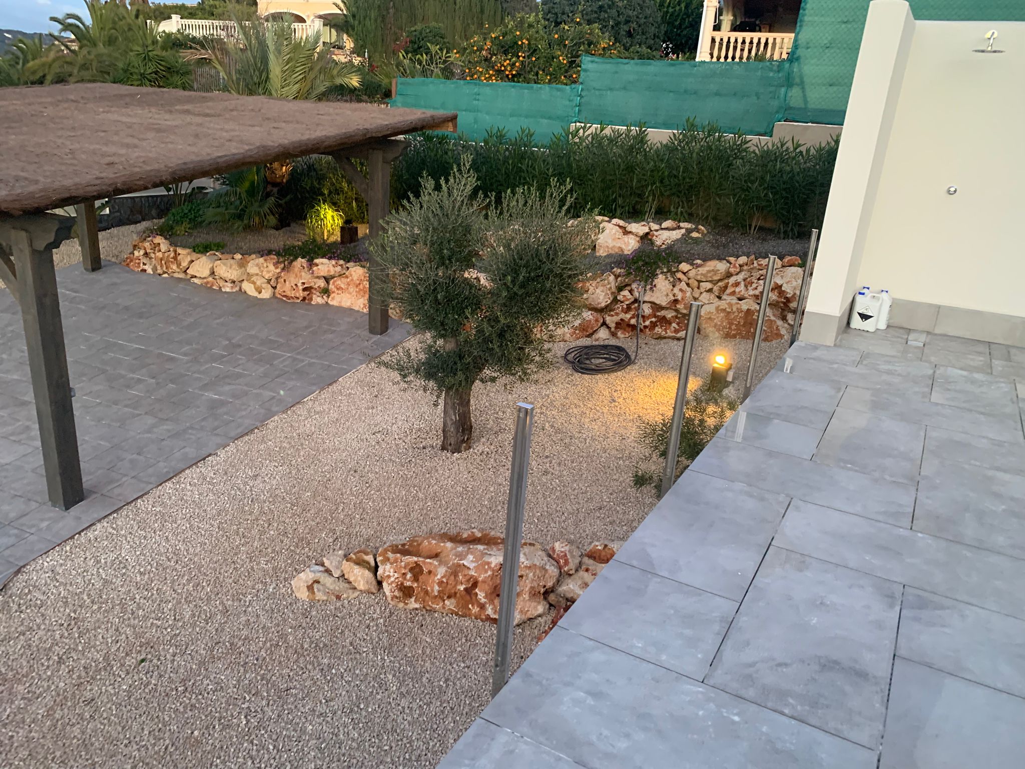 Mediterraner Garten mit Kiesfläche, Olivenbaum, Natursteinbegrenzung, einer Reetdach-Pergola und grauen Steinterrassenfliesen mit Außendusche.