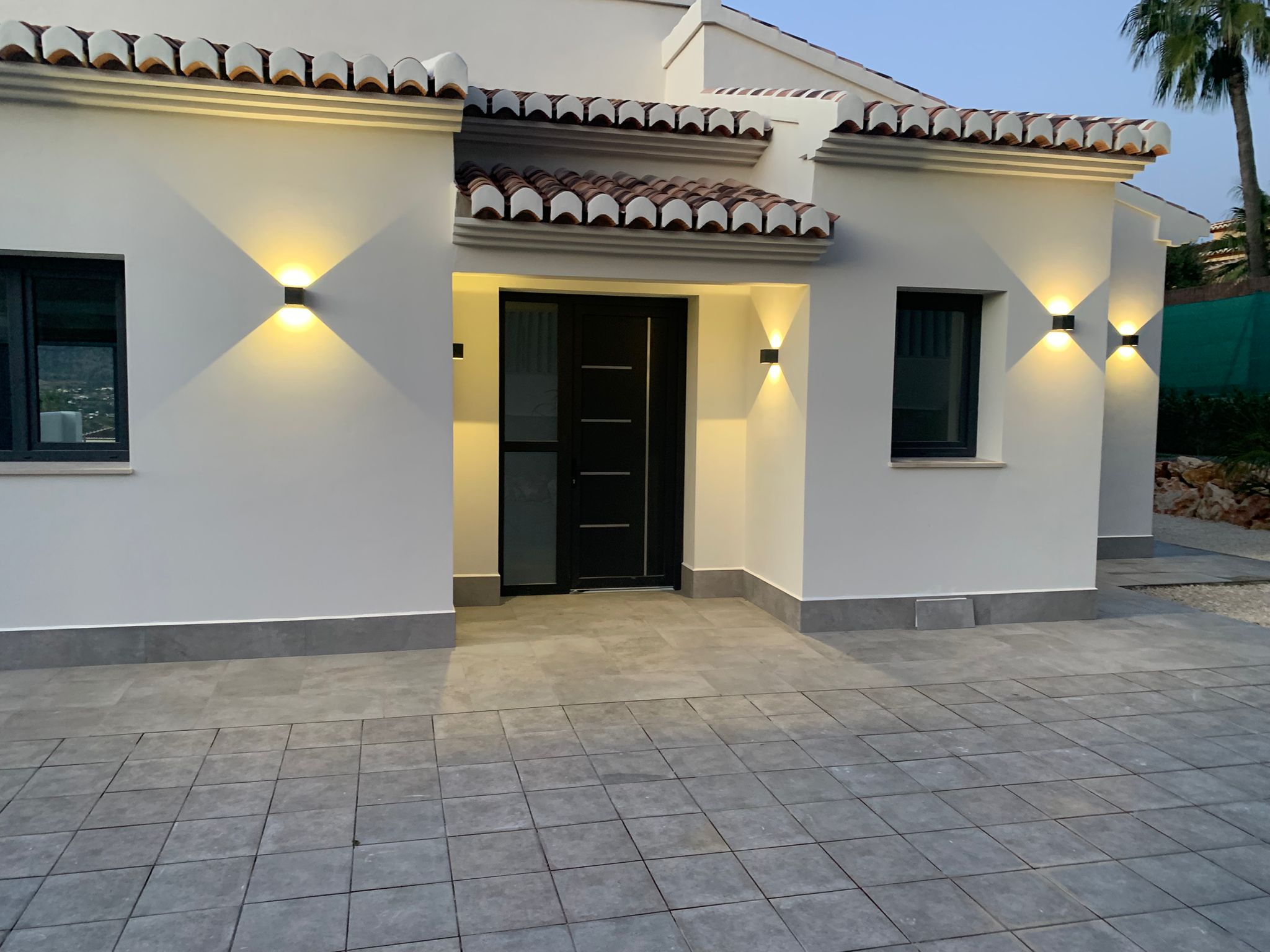 Moderner Villeneingang mit schwarzer Tür, weißer Fassade und grauem Fliesenboden. Symmetrische Wandleuchten bieten warme Beleuchtung unter dem klassischen Ziegeldach.