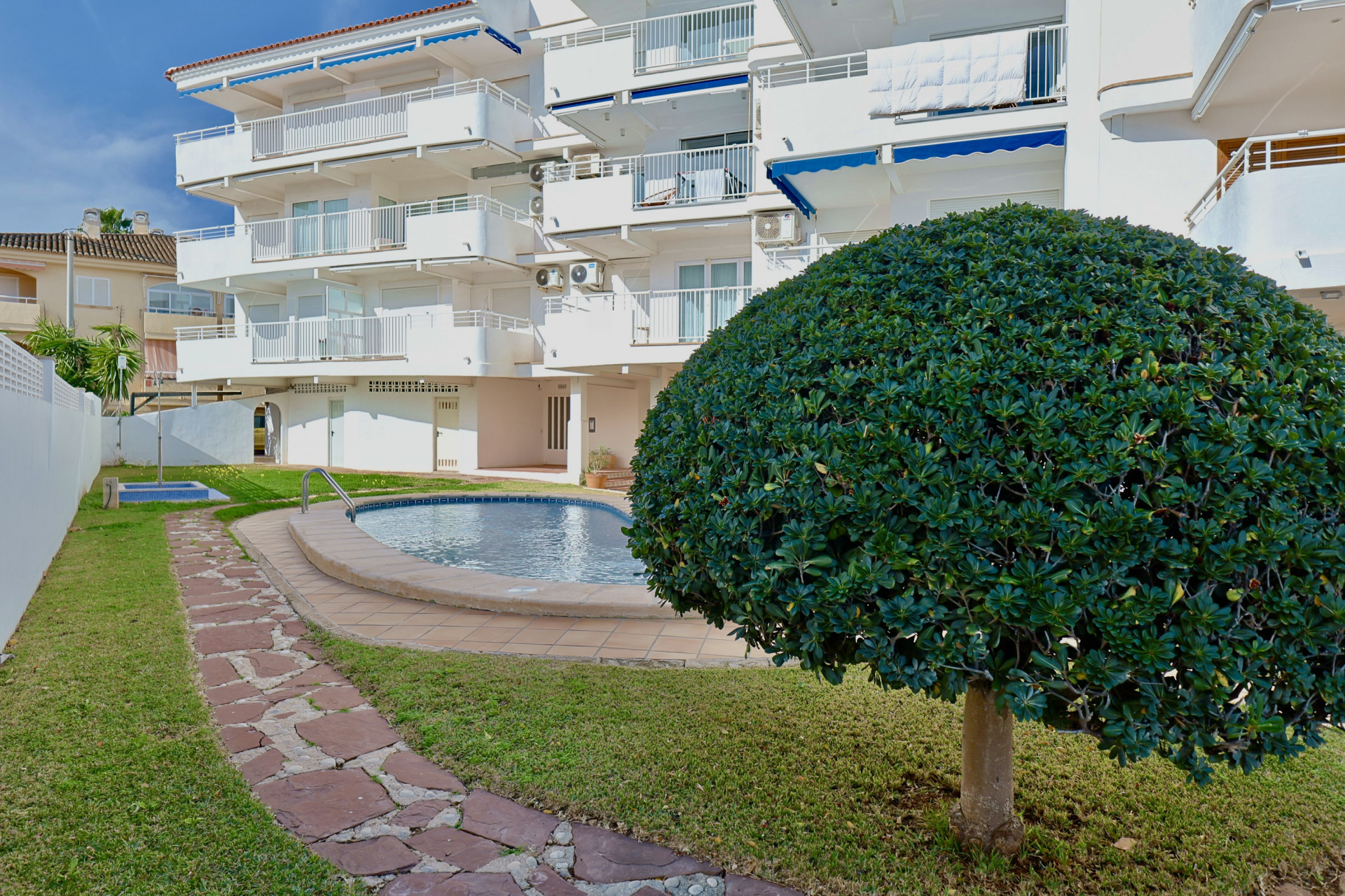 Außenansicht des Apartmentgebäudes mit Gemeinschaftspool, umgeben von grünem Rasen und einem geschwungenen Steinpfad. Ein kugelförmiger Formschnittbusch schmückt den Garten.