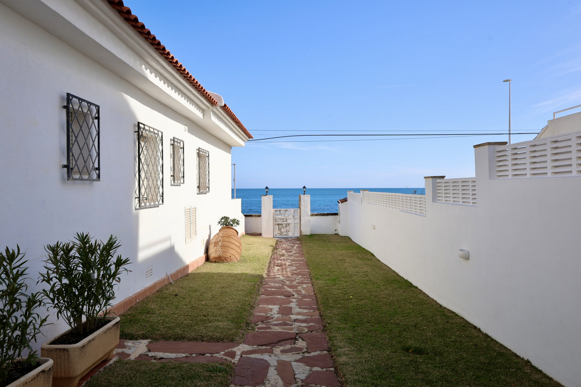 Außenbereich mit Steinplattenweg, der zu einem Tor mit direktem Blick auf das Mittelmeer führt. Das weiße Haus ist mit schmiedeeisernen Fenstergittern und einem Ziegeldach versehen.