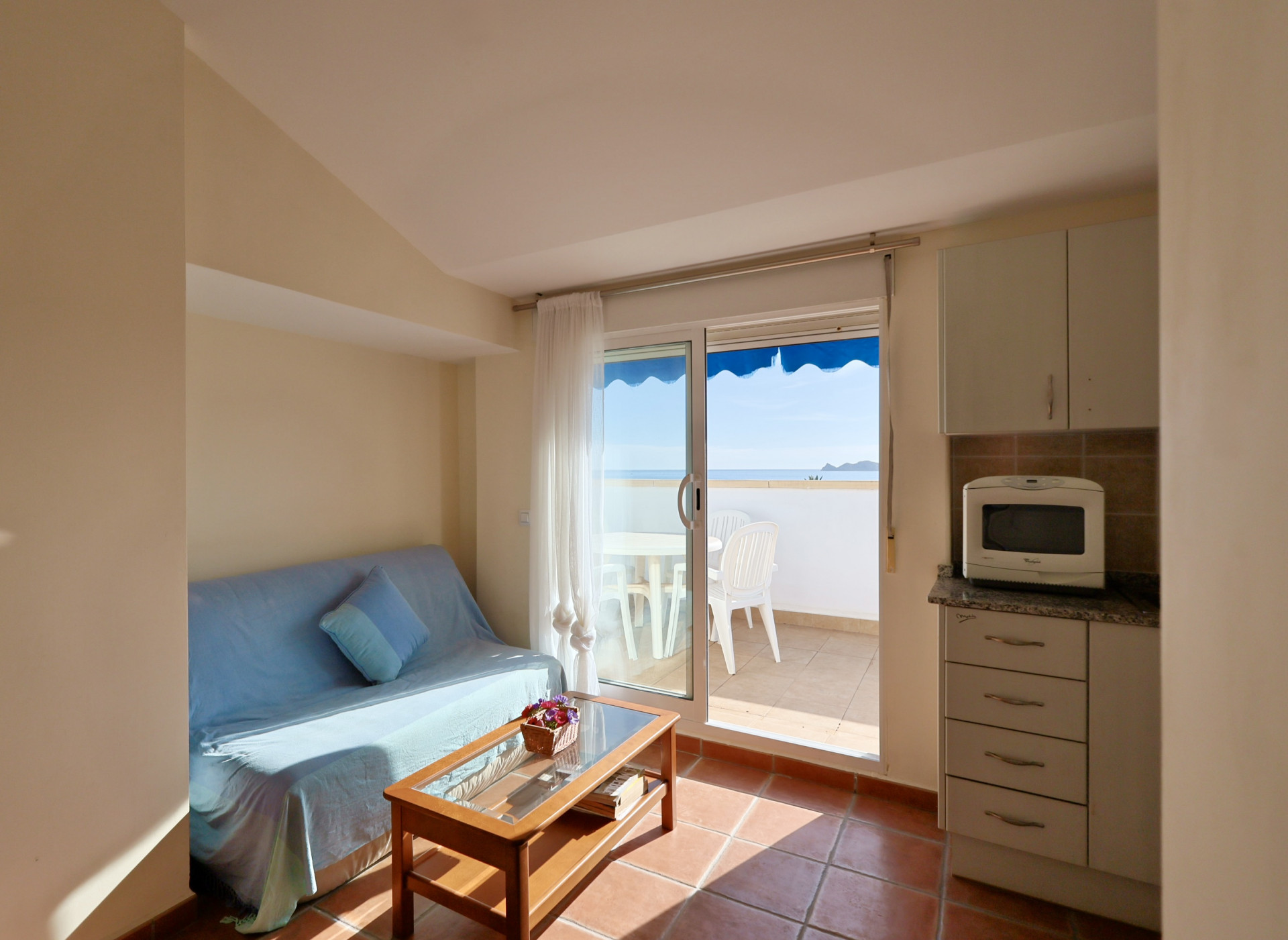 Funktionaler Wohn-Essbereich mit integrierter Küche, Sofa und Couchtisch. Direkter Zugang zu einer gefliesten Terrasse mit Meerblick und Außenmöbeln.