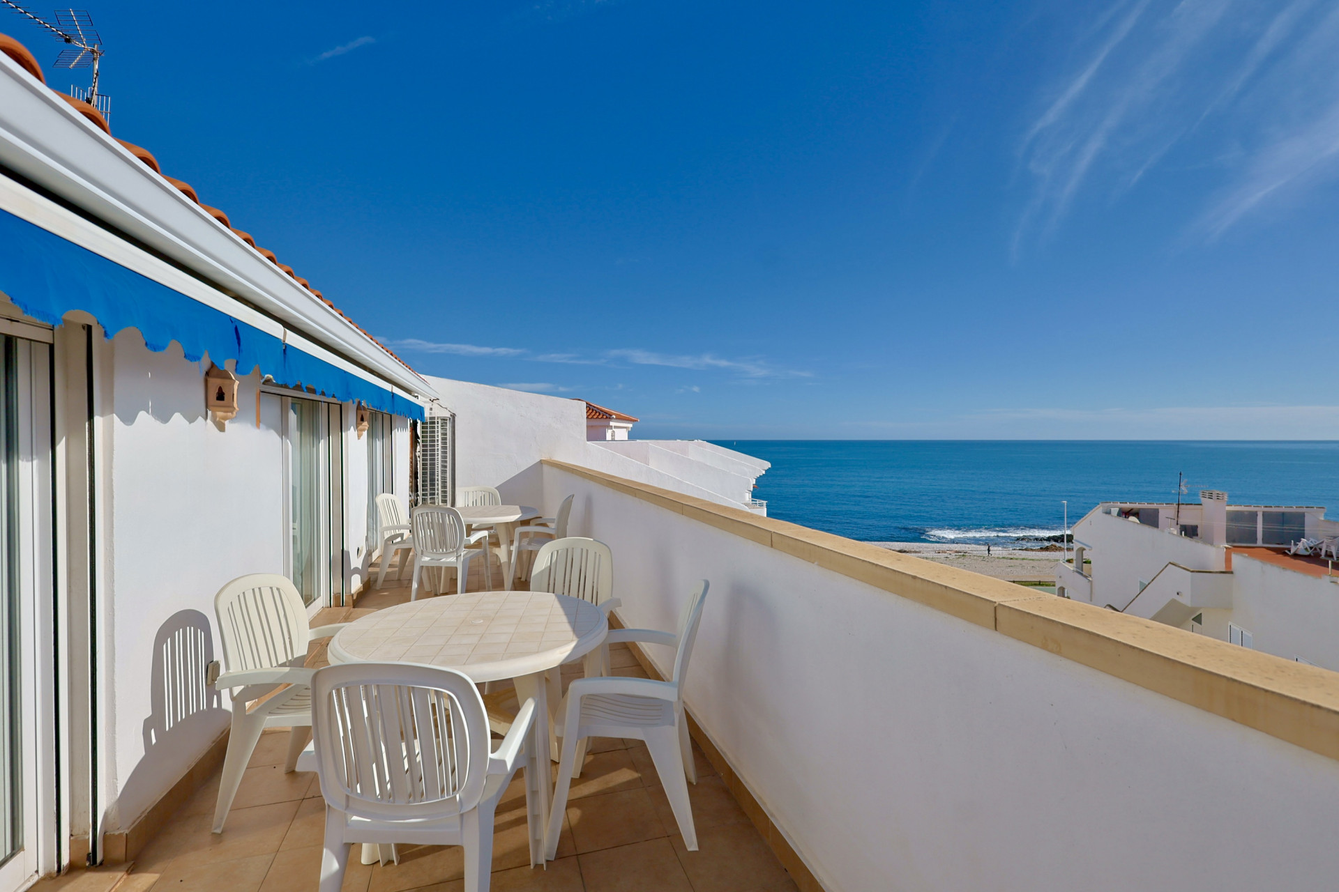 Geräumige private Terrasse mit weißen Gartenmöbeln, blauer Markise und Panoramablick auf das Mittelmeer unter klarem Himmel.