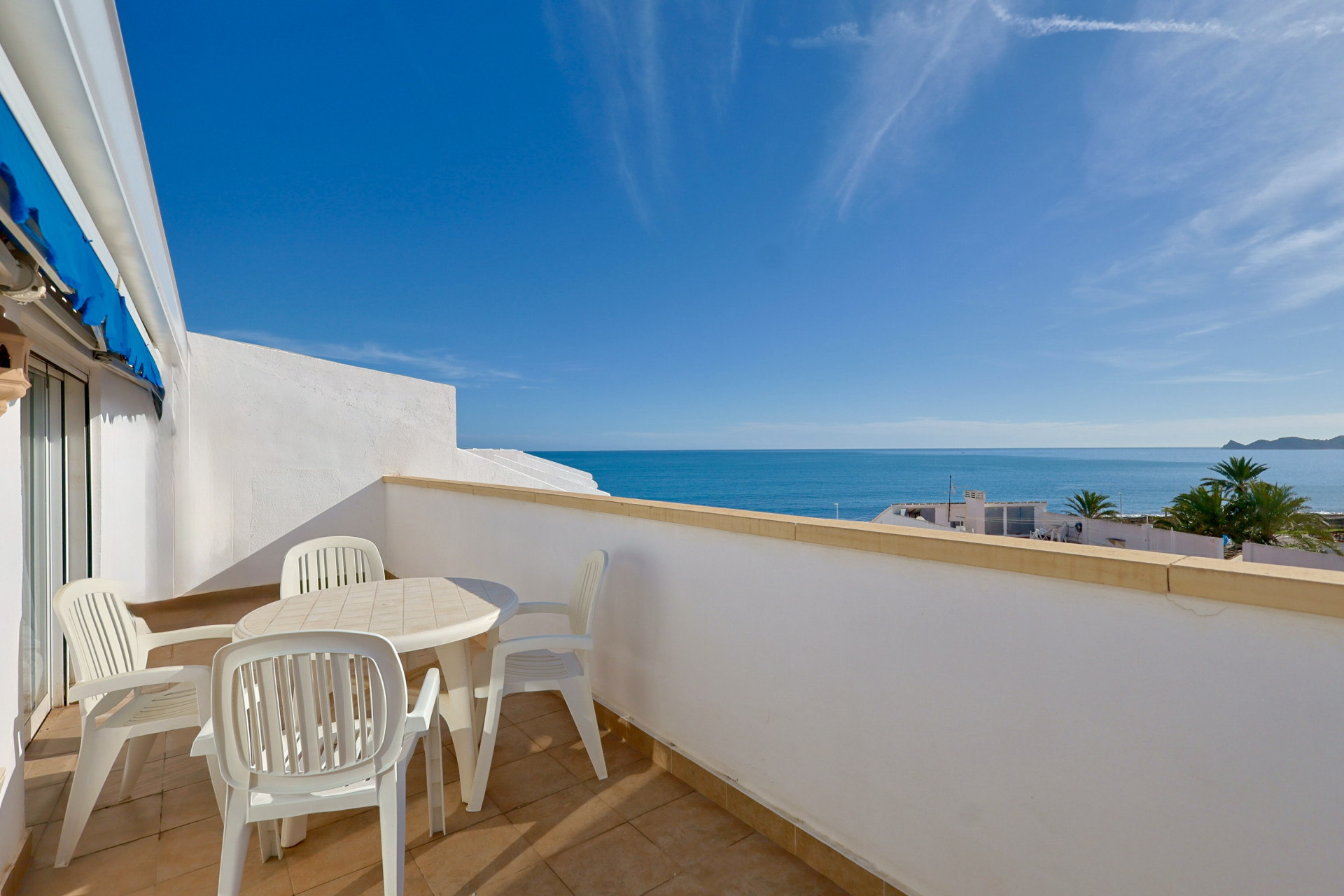 Geräumige Terrasse mit weißen Gartenmöbeln und hellen Fliesen, die einen weiten Blick auf das Mittelmeer und den klaren blauen Himmel bietet. Eine Landzunge ist in der Ferne sichtbar.