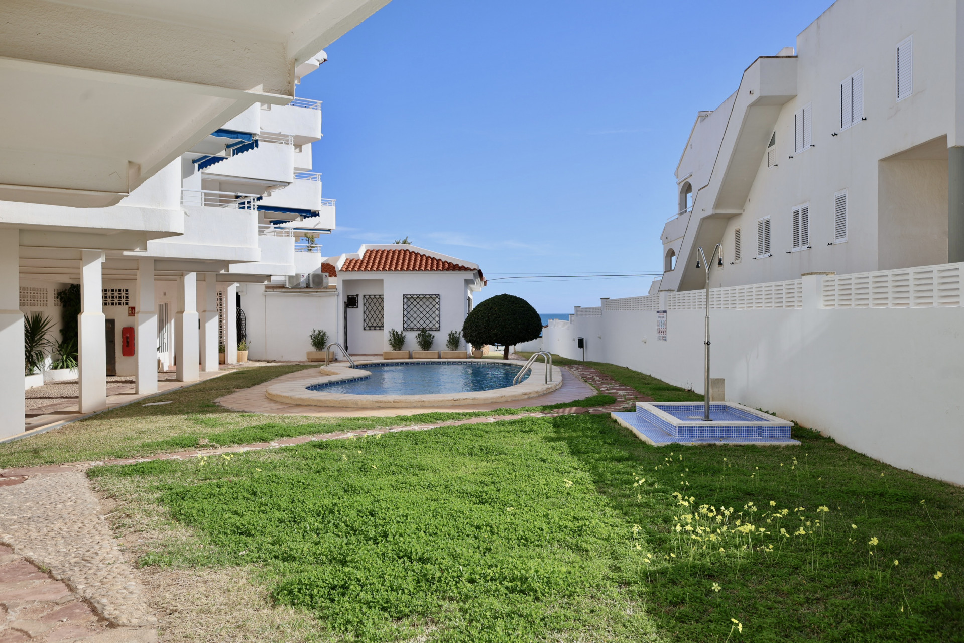 Geschwungener Gemeinschaftspool mit blauen Mosaikfliesen, eingebettet in eine grüne Rasenfläche und weiße mediterrane Gebäude. Das Meer ist im Hintergrund sichtbar.