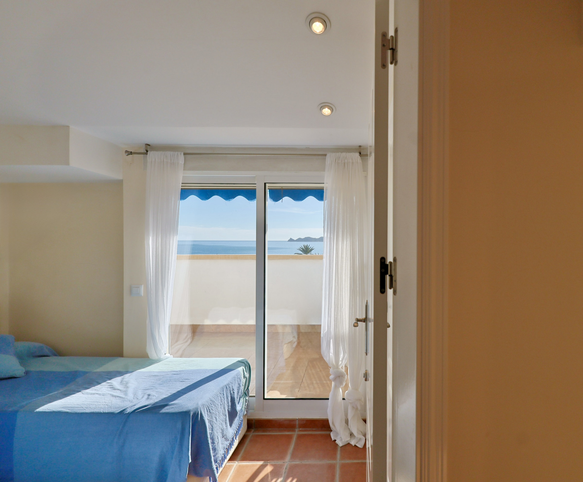 Helles Schlafzimmer mit blau bezogenem Bett und direktem Zugang zu einer Terrasse mit Panoramablick auf das Mittelmeer. Terrakottafliesen und weiße Vorhänge.