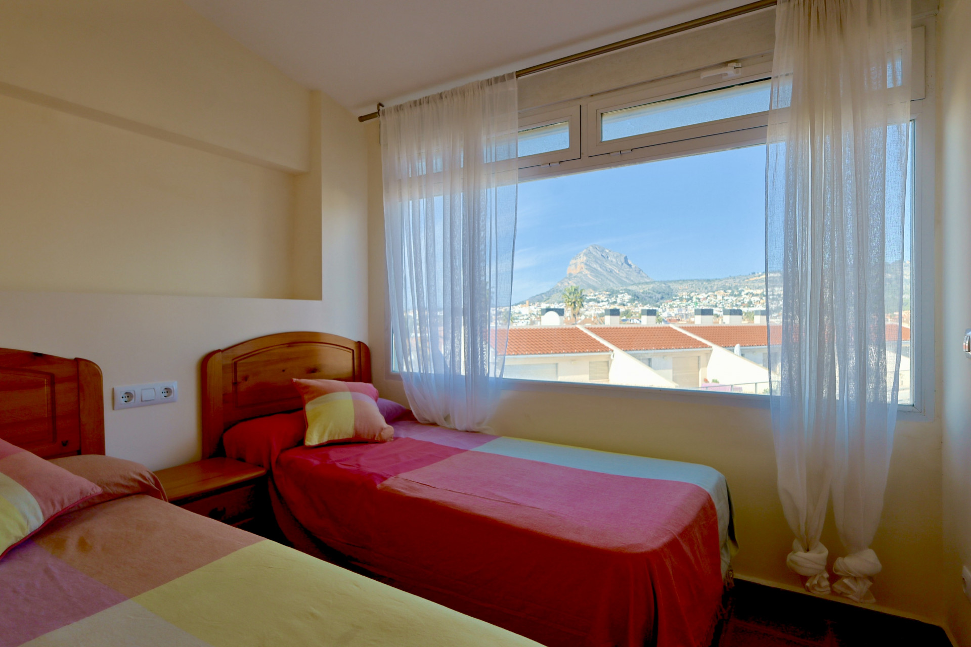 Helles Schlafzimmer mit zwei Einzelbetten und Holz-Kopfteilen, sowie einem großen Fenster mit freiem Blick auf den Berg Montgó und die Dächer.