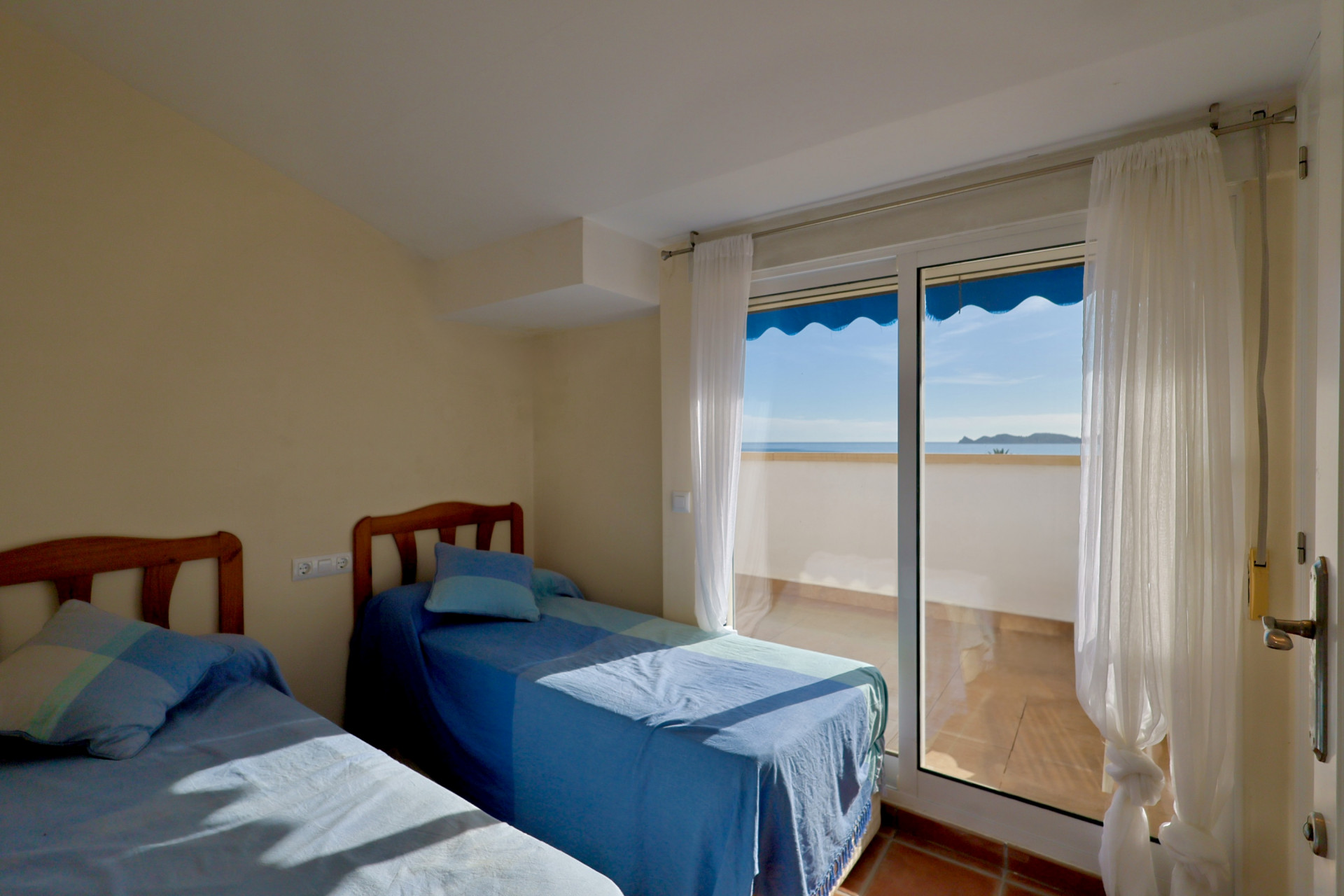 Helles Schlafzimmer mit zwei Einzelbetten und Holz-Kopfteilen. Zugang zu einem Balkon mit Terrakotta-Fliesen, Meerblick und einer blauen Markise.