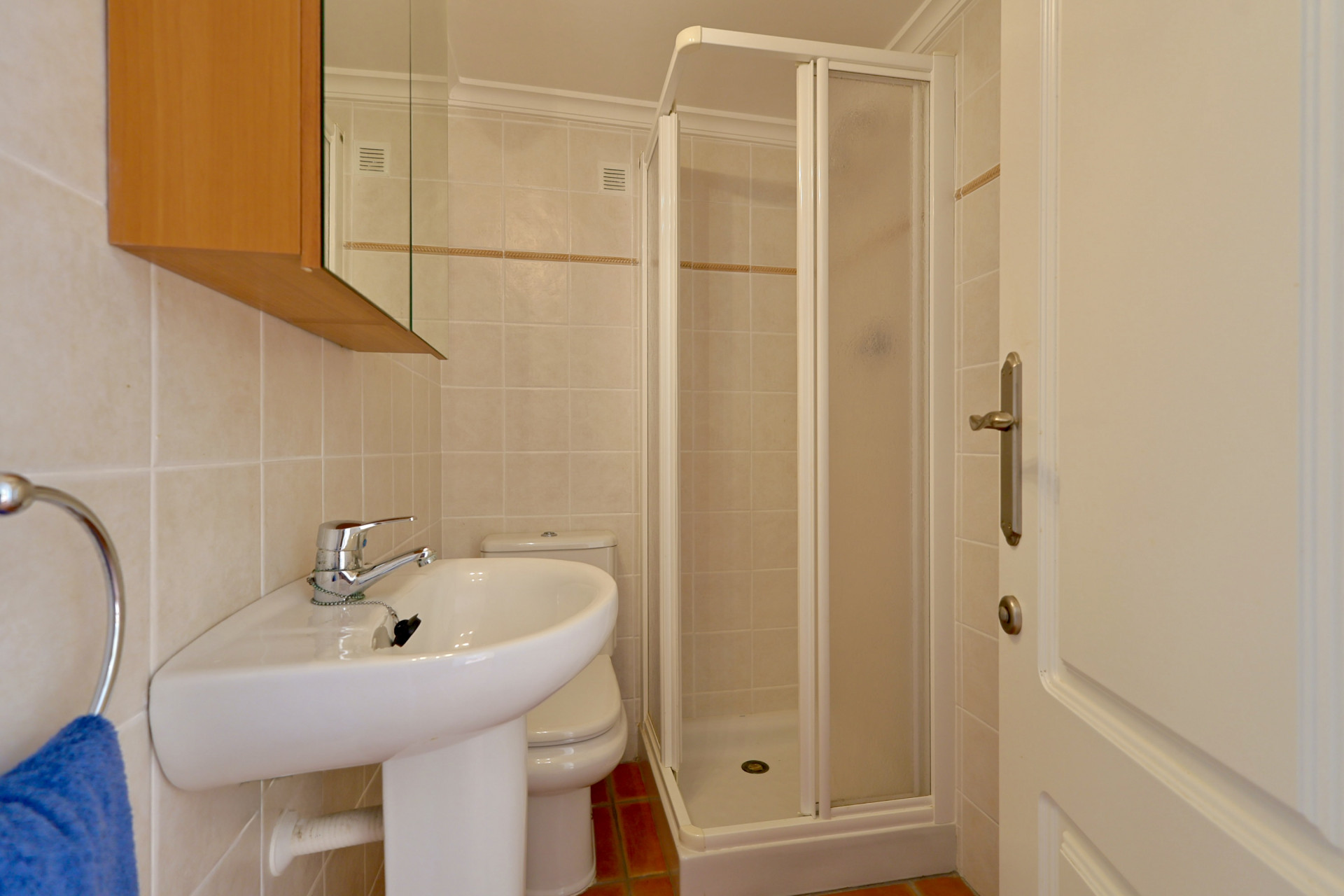 Kompaktes Badezimmer mit Standwaschbecken, WC und Eckduschkabine. Helle Wandfliesen mit Zierleiste und Terrakotta-Bodenfliesen.