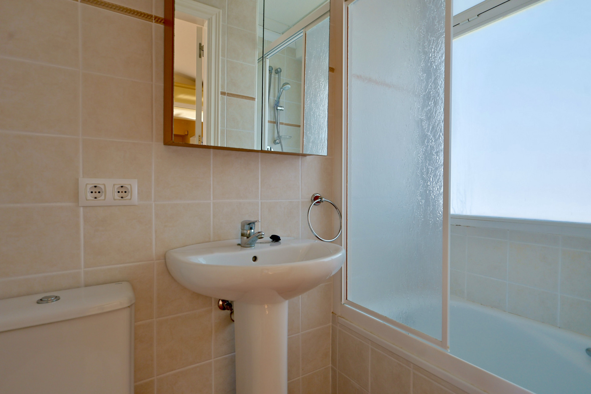 Modernes Badezimmer mit weißem Standwaschbecken, Toilette und hell gefliesten Wänden. Ein Spiegelschrank hängt über dem Waschbecken, neben einer Badewanne mit Milchglasabtrennung.