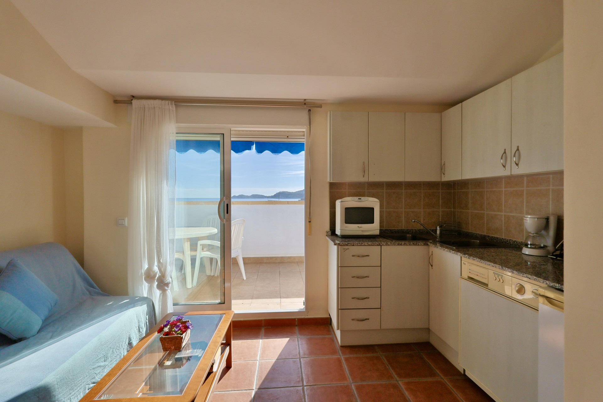 Offener Wohnbereich mit kompakter Küche, weißen Schränken und Terrakotta-Fliesenboden. Eine Glasschiebetür führt zu einem Balkon mit Meerblick und blauer Markise.