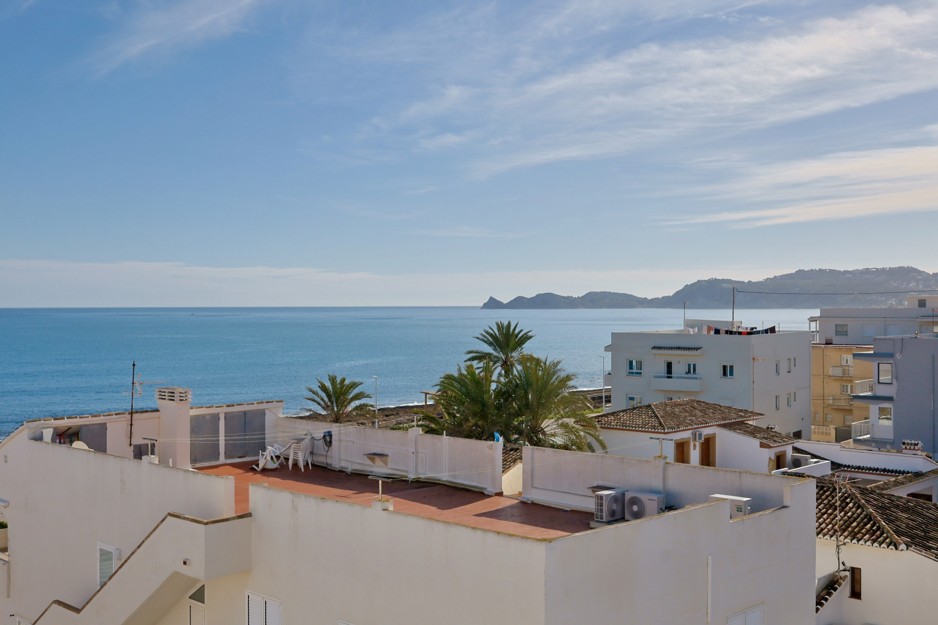 Panoramablick auf das Mittelmeer mit der Küste von Jávea am Horizont, lokale Dächer und Palmen unter einem klaren Himmel.