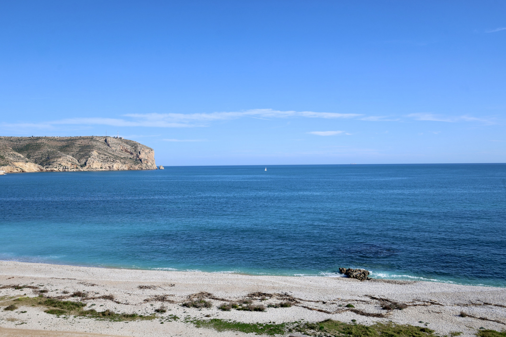 Panoramablick auf einen Kiesstrand mit klarem türkisfarbenem Mittelmeerwasser, einem einzelnen Segelboot am Horizont und einer felsigen Klippe links.