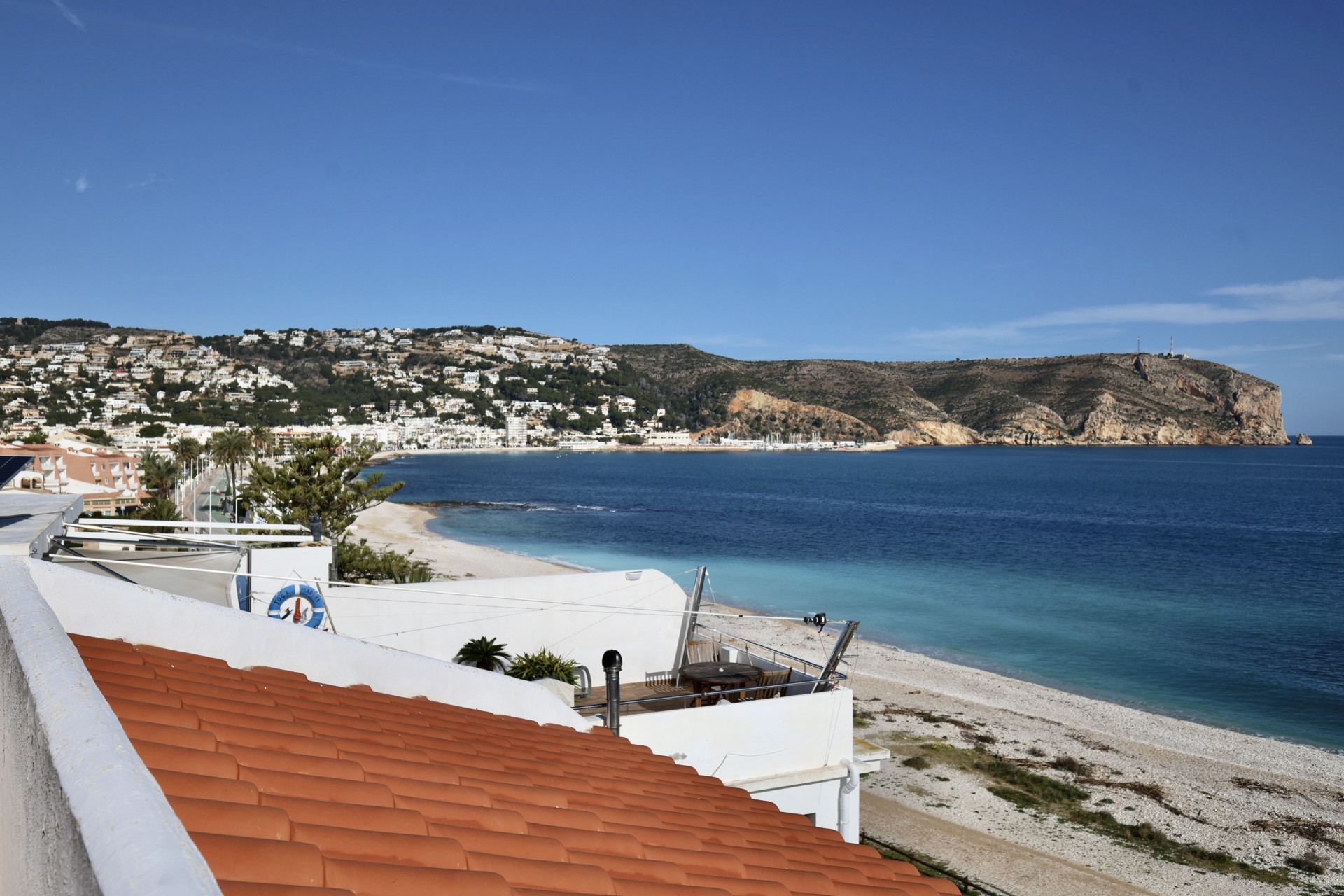 Panoramablick auf Jáveas Küste mit Ziegeldach, Kiesstrand und dem Mittelmeer. Weiße Gebäude am Hang und eine ferne felsige Landzunge sind erkennbar.
