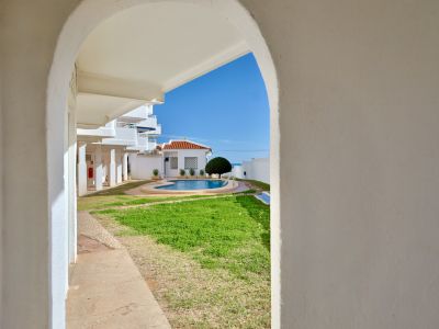 Blick durch einen weißen Bogen auf einen Gemeinschaftspool mit Rasenfläche, ein Gebäude mit Ziegeldach und das Mittelmeer am Horizont unter blauem Himmel.