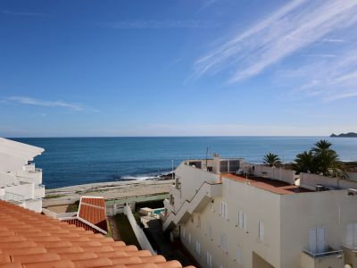 Dachansicht mit Terrakotta-Ziegeln, die einen Panoramablick auf das Mittelmeer und einen Kiesstrand bietet. Weiße Küstengebäude und eine ferne Landzunge unter klarem blauem Himmel.