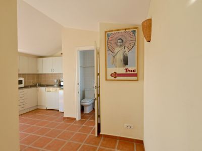 Flur mit Terrakottafliesen, der zu einer offenen Küche mit weißen Schränken und einem Badezimmerzugang führt. Ein gerahmtes 'Thai'-Poster schmückt die Wand.