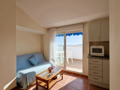 Funktionaler Wohn-Essbereich mit integrierter Küche, Sofa und Couchtisch. Direkter Zugang zu einer gefliesten Terrasse mit Meerblick und Außenmöbeln.