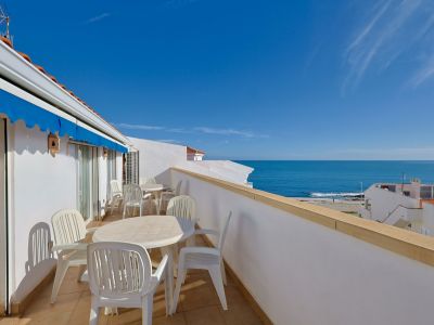 Geräumige private Terrasse mit weißen Gartenmöbeln, blauer Markise und Panoramablick auf das Mittelmeer unter klarem Himmel.