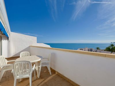 Geräumige Terrasse mit weißen Gartenmöbeln und hellen Fliesen, die einen weiten Blick auf das Mittelmeer und den klaren blauen Himmel bietet. Eine Landzunge ist in der Ferne sichtbar.