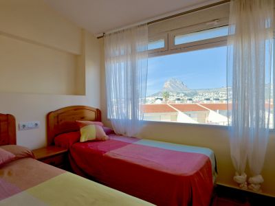 Helles Schlafzimmer mit zwei Einzelbetten und Holz-Kopfteilen, sowie einem großen Fenster mit freiem Blick auf den Berg Montgó und die Dächer.