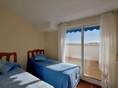 Helles Schlafzimmer mit zwei Einzelbetten und Holz-Kopfteilen. Zugang zu einem Balkon mit Terrakotta-Fliesen, Meerblick und einer blauen Markise.