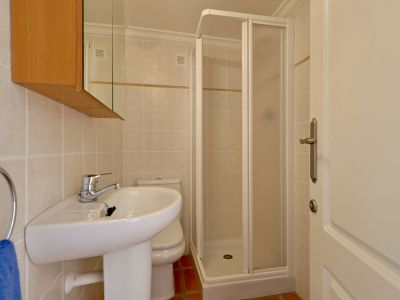 Kompaktes Badezimmer mit Standwaschbecken, WC und Eckduschkabine. Helle Wandfliesen mit Zierleiste und Terrakotta-Bodenfliesen.