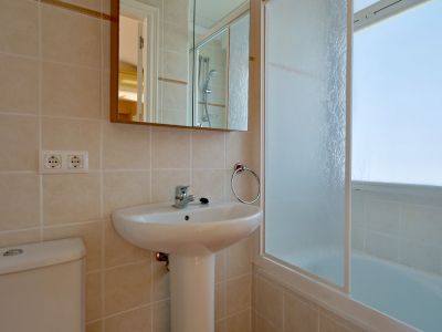 Modernes Badezimmer mit weißem Standwaschbecken, Toilette und hell gefliesten Wänden. Ein Spiegelschrank hängt über dem Waschbecken, neben einer Badewanne mit Milchglasabtrennung.