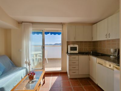 Offener Wohnbereich mit kompakter Küche, weißen Schränken und Terrakotta-Fliesenboden. Eine Glasschiebetür führt zu einem Balkon mit Meerblick und blauer Markise.