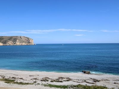 Panoramablick auf einen Kiesstrand mit klarem türkisfarbenem Mittelmeerwasser, einem einzelnen Segelboot am Horizont und einer felsigen Klippe links.