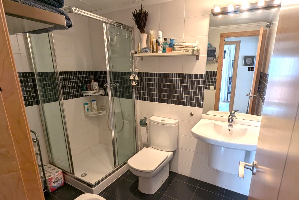 Badezimmer mit Glasduschkabine, weißen Wandfliesen mit dunkler Mosaikbordüre und anthrazitfarbenem Bodenbelag.