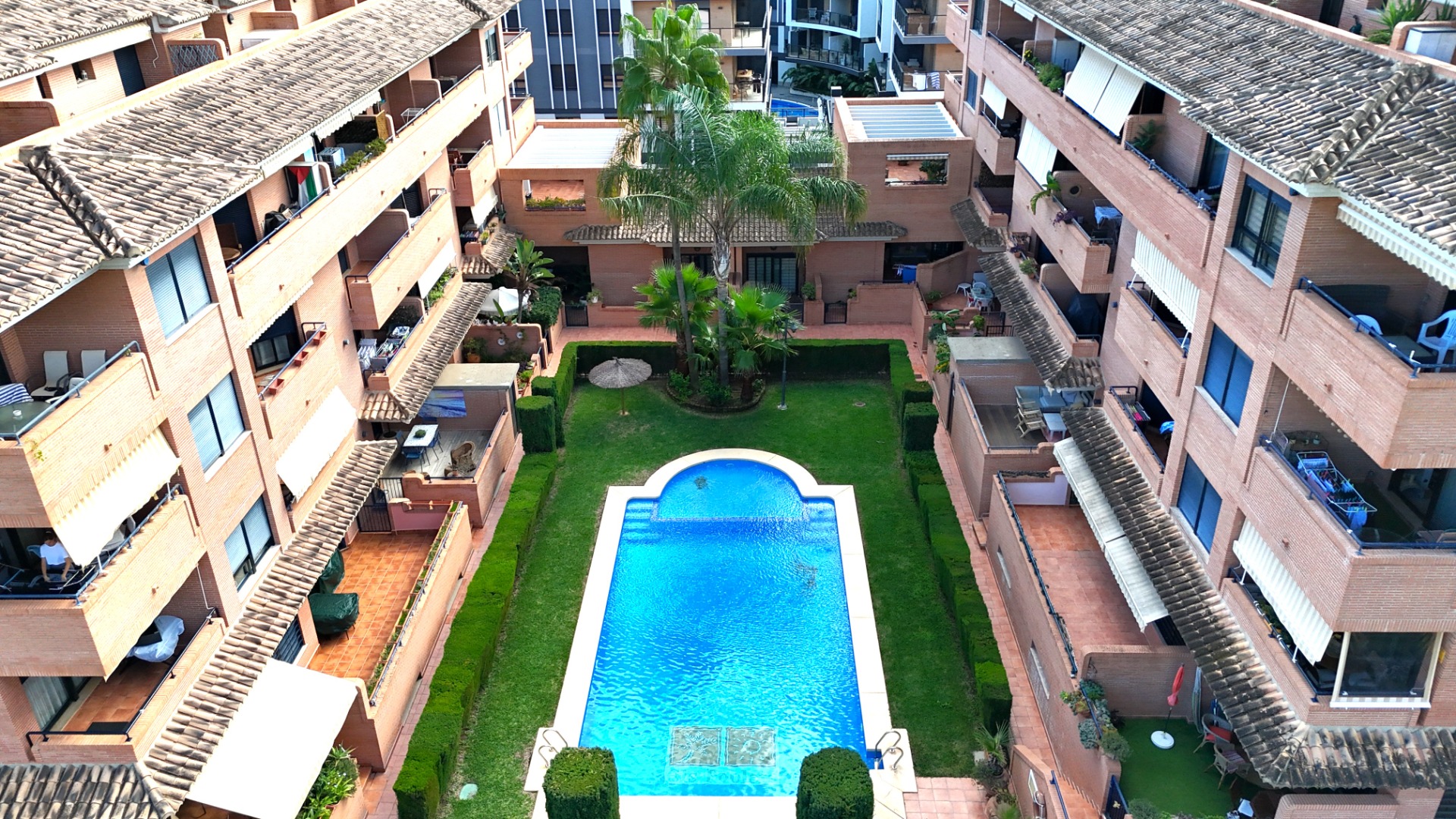 Erhöhte Ansicht des Gemeinschaftsbereichs mit Pool, gepflegtem Rasen und Palmen, umgeben von Backsteingebäuden mit privaten Terrassen in Jávea.