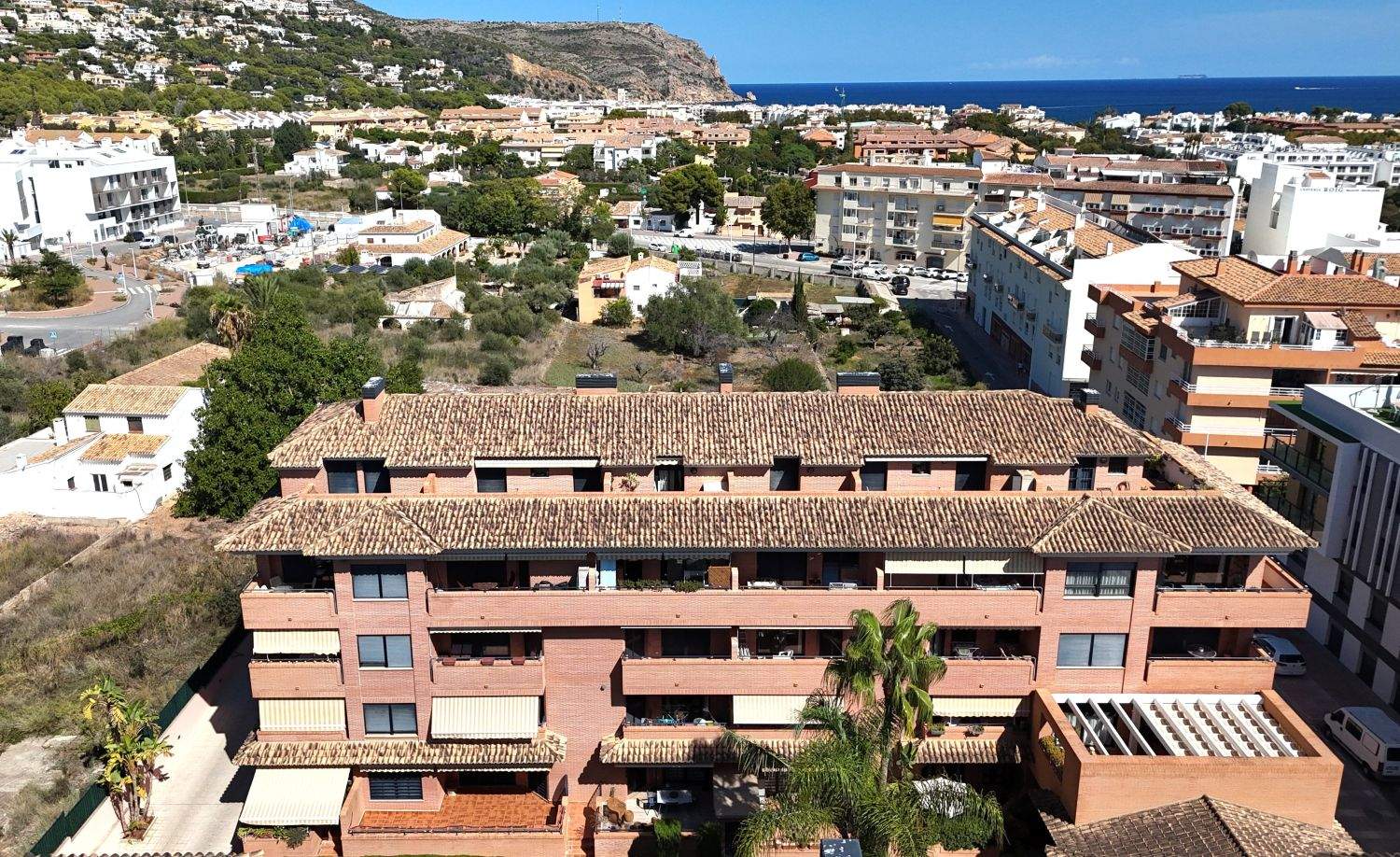 Erhöhte Ansicht eines Backsteingebäudes mit Terrakottadächern, Balkonen mit Markisen und weitem Blick über Jávea bis hin zum Mittelmeer.
