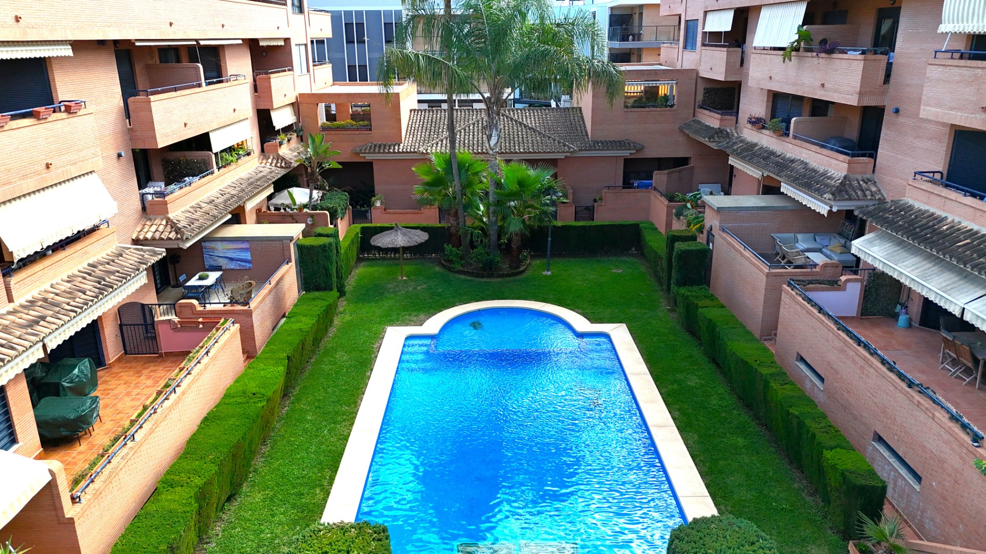 Gemeinschaftsanlage mit rechteckigem Pool, gepflegter Rasenfläche und Palmen, umgeben von Backsteingebäuden mit privaten Terrassen.