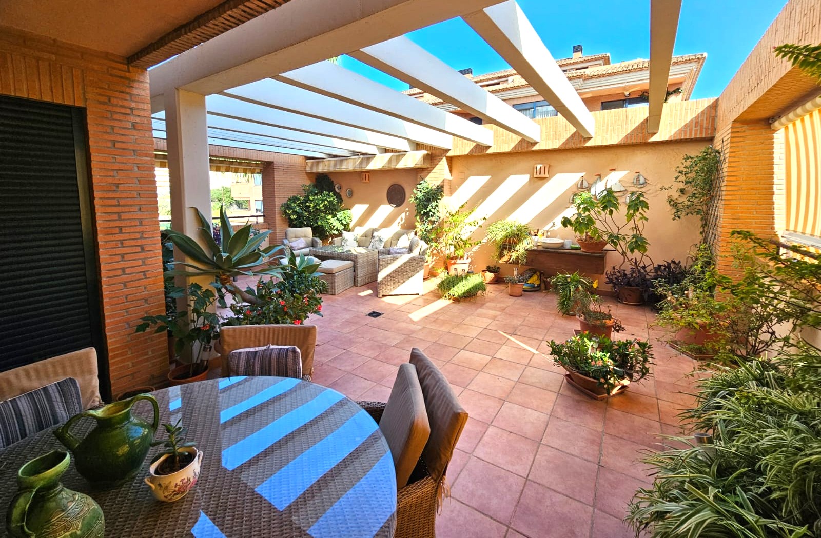 Großzügige mediterrane Terrasse mit Terrakottaboden, weißer Pergola und kombinierten Ess- und Lounge-Bereichen inmitten üppiger Topfpflanzen.
