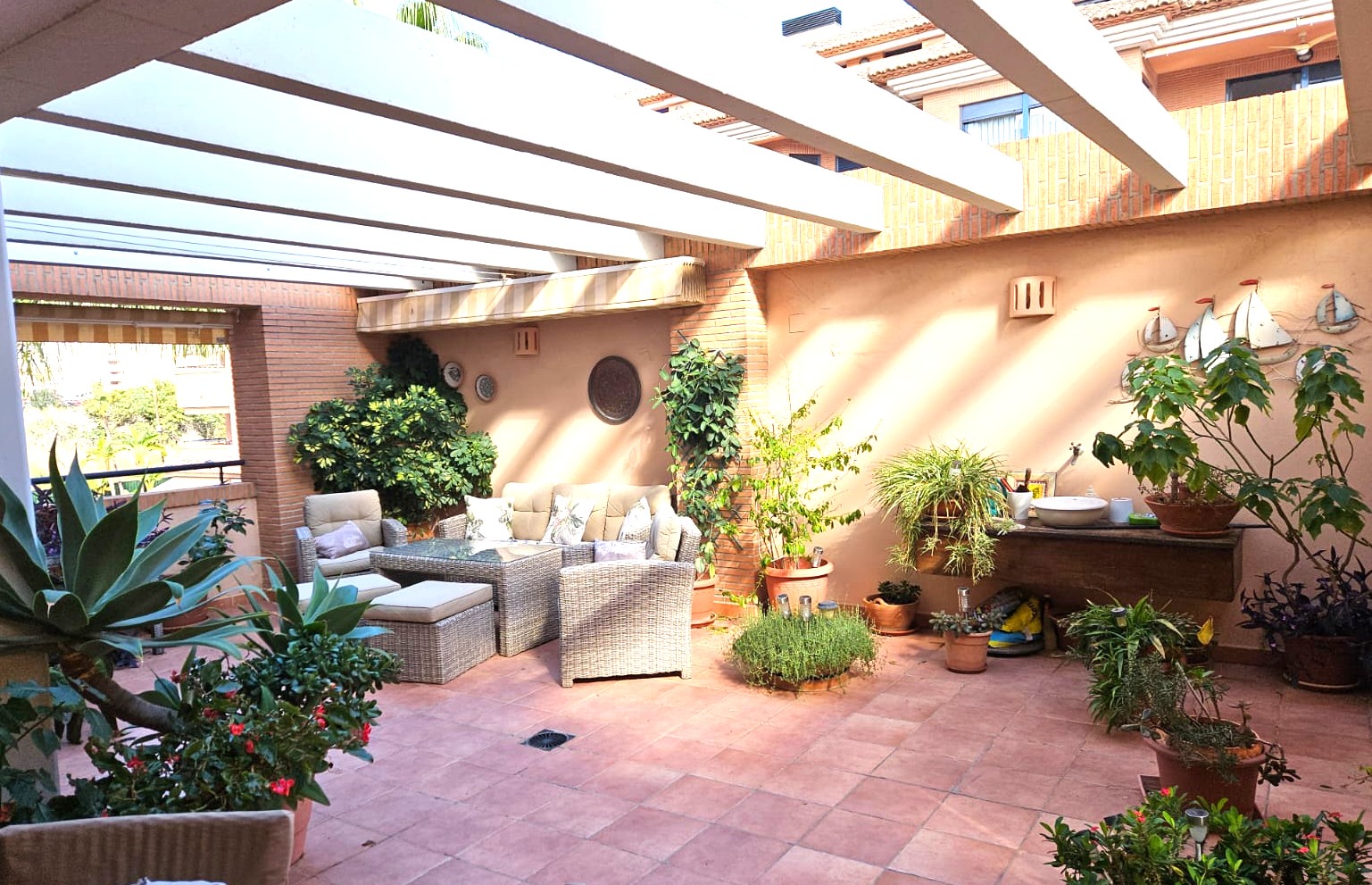 Großzügige private Terrasse mit Terrakottaboden und weißen Pergola-Balken. Ausgestattet mit Rattan-Loungemöbeln, Backsteinwänden und vielfältiger mediterraner Bepflanzung.