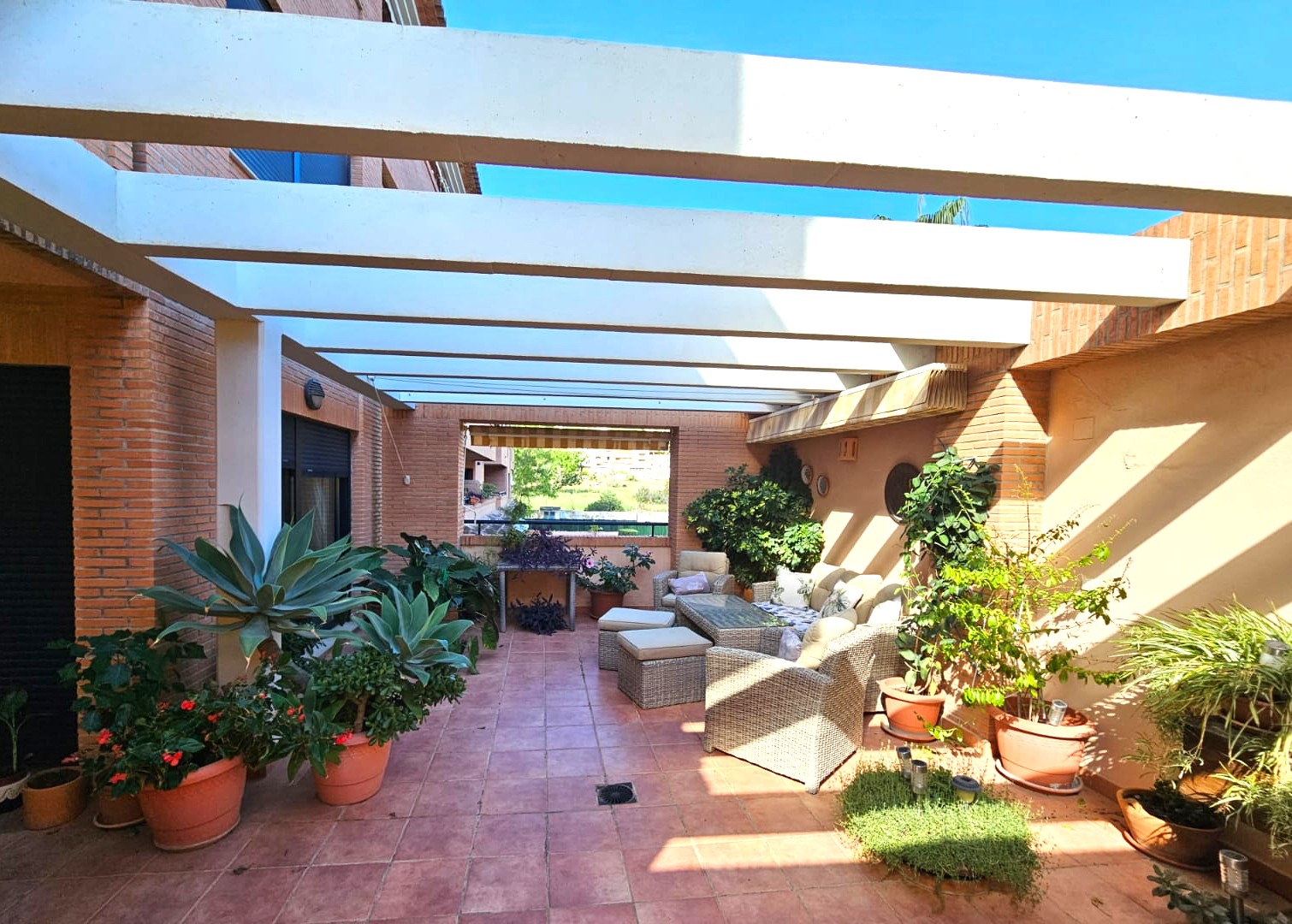 Großzügige Terrasse mit Terrakottaboden, weißen Pergola-Balken und einer Rattan-Sitzgruppe, umgeben von mediterranen Topfpflanzen und Backsteinwänden.