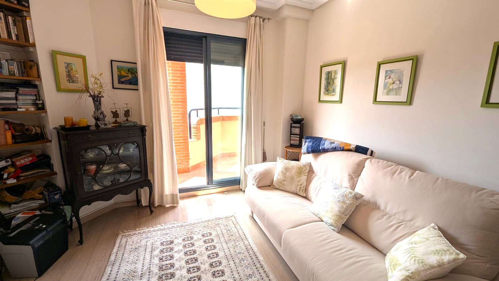 Heller Wohnbereich mit beigem Sofa, antikem dunklem Holzvitrinenschrank und Glasschiebetüren, die auf einen Balkon mit Backsteinmauer führen.