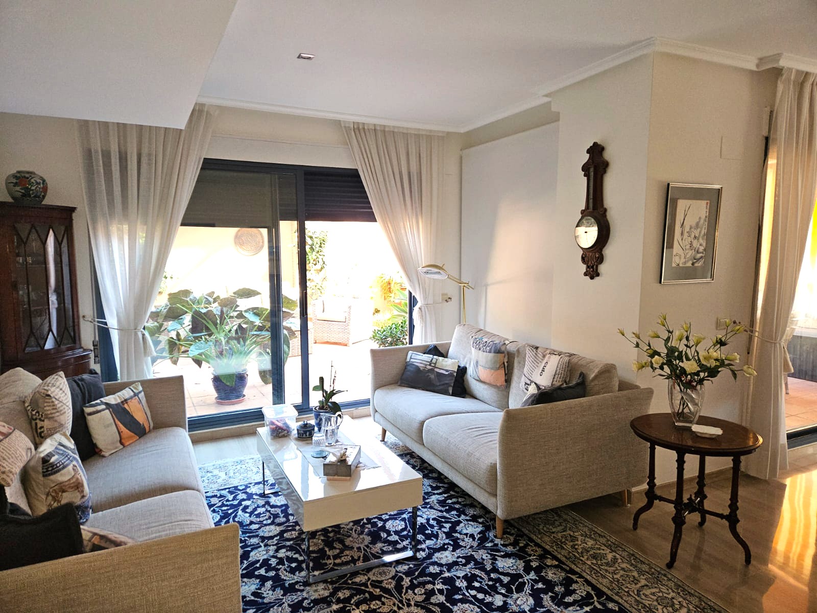 Heller Wohnbereich mit zwei neutralen Sofas, gemustertem blauem Teppich und großen Glasschiebetüren, die auf eine private Terrasse im Grünen führen.