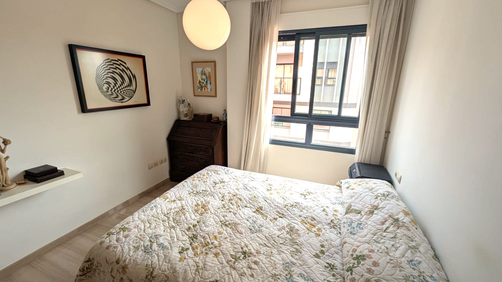 Helles Schlafzimmer mit antikem Holzsekretär, großem Fenster mit hellen Vorhängen und einer markanten kugelförmigen Deckenleuchte über dem Bett.